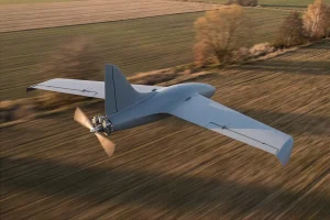 Rear view of the MTS autonomous combat UAV in flight, showing its pusher propeller and tail section over a field.